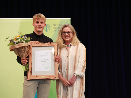Naturschutzpreis-Gewinner Matteo Schölzel steht mit Blumenstrauß und Urkunde neben Umweltsenatorin Ute Bonde auf einer Bühne 