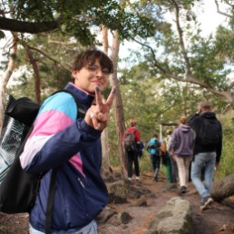 Ein junger Mensch zeigt das Victory-Zeichen während er auf einem Waldweg läuft. Vor ihm sind weitere Menschen zu sehen. 